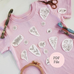 May include: A light pink t-shirt decorated with various white fabric patches featuring black line art of flowers, leaves, and a bee. The shirt is accompanied by embroidery floss, a pair of metal scissors, and a wooden embroidery hoop.