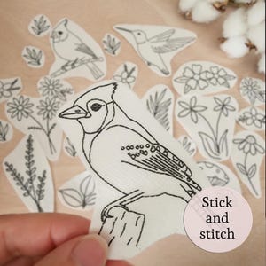 May include: A collection of white fabric patches with black embroidered designs of birds, flowers, and leaves. The patches include a blue jay, hummingbird, and other floral elements. A pink circle with the words "Stick and stitch" is in the lower right corner.