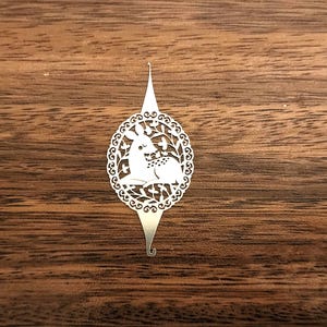以下が含まれることがあります： 銀色の装飾的なブックマーク。花輪の中にいる鹿の切り絵のデザインが特徴です。 ブックマークは、尖った上部と下部に湾曲したフックが付いています。 背景は濃い木材の表面です。