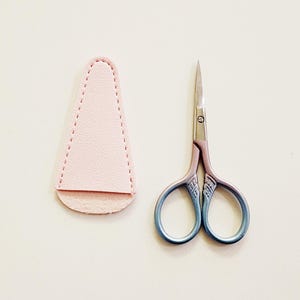 May include: A pair of small, decorative scissors with a blue and purple handle, next to a pink leather sheath. The scissors have a pointed tip and are designed for detailed cutting tasks.