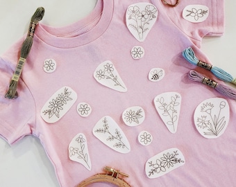 Puede incluir: Una camiseta rosa claro decorada con varios parches de tela blanca con dibujos de líneas negras de flores, hojas y una abeja. La camiseta está rodeada de artículos de bordado, incluyendo un bastidor de madera, tijeras de cobre y tres madejas de hilo.
