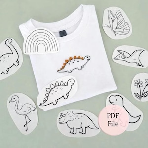 May include: A white t-shirt featuring an embroidered dinosaur with orange spikes, surrounded by various iron-on patches including a rainbow, a butterfly, a pterodactyl, a flamingo, a stegosaurus, a triceratops, a flower, and a PDF File label.