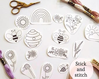 May include: A selection of white fabric patches featuring black line art designs, such as bees, flowers, a rainbow, and a beehive. Gold scissors, embroidery threads, and a wooden hoop are also visible. The text "Stick and stitch" is in a pink circle.