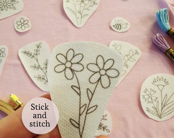 Puede incluir: Una colección de parches estabilizadores de bordado adhesivos blancos con dibujos de líneas negras de flores, hojas y una abeja. Una pegatina circular con el texto Stick and stitch es visible junto a hilo de bordar azul y morado.