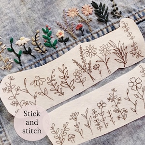 May include: Two off-white fabric strips with brown floral line art, reading "Stick and stitch". The strips are placed on a denim jacket with embroidered flowers in various colors. The floral designs include daisies, leaves, and other botanical elements.