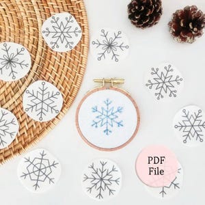 May include: A wooden embroidery hoop containing white fabric with a blue stitched snowflake design, surrounded by several white paper cutouts featuring various gray snowflake illustrations and two brown pinecones with glitter accents.