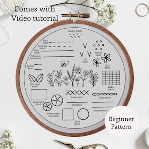May include: A round embroidery hoop with a light gray fabric featuring various stitch types, floral designs, and the text "Beginner Pattern." The text "Comes with Video tutorial" is also visible. The hoop is framed in wood.