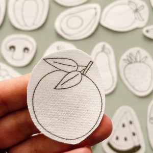 May include: A white felt patch shaped like a round orange with two leaves, featuring a black embroidered outline. The patch is part of a collection of various fruit and vegetable shapes, each with a similar black stitched border design.