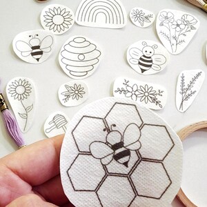 May include: A collection of white fabric patches featuring black line art designs including bees, beehives, honeycombs, sunflowers, and floral arrangements. The patches are arranged alongside a wooden embroidery hoop and lavender embroidery floss.