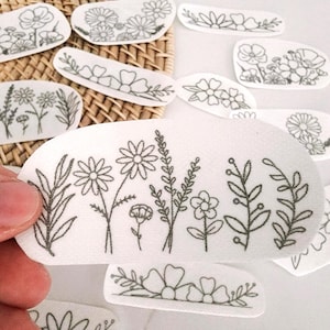 May include: A collection of white adhesive patches featuring black line art illustrations of various wildflowers, daisies, and leafy stems. Each patch displays a unique botanical arrangement with thin, detailed outlines of floral elements.