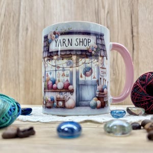 Könnte beinhalten: Eine Keramiktasse mit rosa Griff zeigt eine Aquarellillustration eines Wollgeschäfts. Das Schild des Geschäfts lautet "YARN SHOP" über einer detaillierten Fassade mit Wollknäueln und Körben. Ein Knäuel blaue Wolle und dekorative Steine befinden sich im Vordergrund.