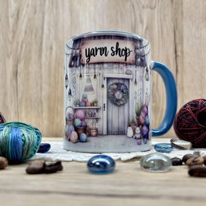Könnte beinhalten: Eine Keramik-Tasse mit blauem Henkel und Rand, mit einer Aquarell-Illustration eines Wollgeschäfts. Das Geschäft hat eine Holzfassade, einen Kranz an der Tür und ist mit Wollknäueln gefüllt. Der Text "yarn shop" steht darüber.