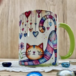 Könnte beinhalten: Eine weiße Keramiktasse mit einer farbenfrohen Illustration einer sich streckenden Katze. Die Katze hat orange-braunes Fell mit einem gestreiften Schwanz. Die Tasse zeigt hängende Herzen und einen grünen Henkel. Dekorative Glasperlen sind um die Tasse verstreut.