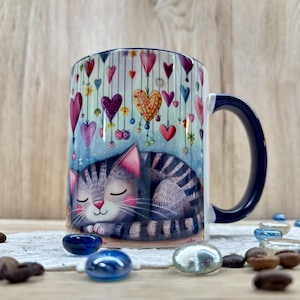 Könnte beinhalten: Eine Keramiktasse mit marineblauem Griff und Rand. Die Tasse zeigt eine Illustration einer schlafenden grauen Tabby-Katze mit rosa Wangen. Die Tasse ist mit hängenden Herzen in verschiedenen Farben und Mustern vor blauem Hintergrund verziert.