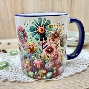 Könnte beinhalten: Weiße Keramiktasse mit einem farbenfrohen Blumendesign. Die Tasse hat einen blauen Henkel und Rand. Das Blumenmuster zeigt skurrile Blumen in Rosa, Blau, Grün und Gelb.