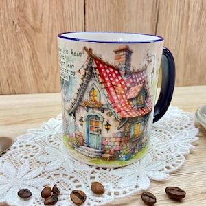 Könnte beinhalten: Eine weiße Keramiktasse mit blauem Rand und Henkel. Die Tasse zeigt eine farbenfrohe Illustration eines fantasievollen Häuschens mit einem rot-weiß gepunkteten Dach. Der Text auf der Tasse lautet "Liebe ist kein Ort, sondern ein Gefühl!"