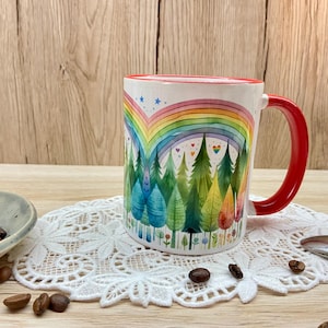 Könnte beinhalten: Weiße Keramik-Tasse mit rotem Henkel und Rand, mit einem bunten Regenbogen- und Wald-Design. Die Tasse steht auf einer weißen Spitzendecke, mit Kaffeebohnen und einer kleinen Schale in der Nähe.