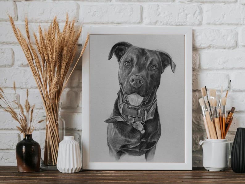 May include: A framed charcoal drawing of a dog wearing a collar and bandana. The dog is depicted with its mouth open and tongue visible. The artwork is presented in a white frame, positioned on a surface with decorative dried wheat stalks.