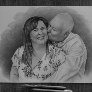 May include: A graphite pencil drawing on paper depicting a smiling person with shoulder-length hair wearing a floral print top, being kissed on the cheek by a bald person wearing a collared shirt. The artwork features detailed shading.