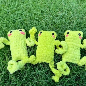 May include: Three handmade, light green crochet frog toys with black eyes and pink cheeks are displayed on a bed of green grass. The frogs have outstretched arms and legs, giving them a playful appearance.