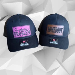 May include: Two black trucker hats with mesh back panels. One hat has a pink patch with the text "Somebody's Problem" in black. The other hat has a brown patch with the text "Somebody" in black.