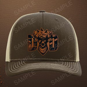 May include: A dark olive green baseball cap with a mesh back and a front graphic featuring the word DAD with a deer head silhouette integrated into the center letter. The design includes a camouflage pattern and small bird silhouettes above the deer.