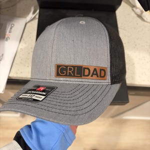 May include: A grey and black trucker hat featuring a rectangular brown leather patch on the front with the text GRL DAD printed in black. The hat has a structured grey front panel, a black mesh back, and a sticker on the brim reading Richardson 112.