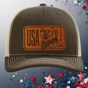 May include: A brown and tan trucker hat featuring a rectangular patch with an orange graphic of an eagle, the text USA, 1776, and LIVE FREE. The hat has a structured front panel, a mesh back, and contrast stitching along the brim and crown.