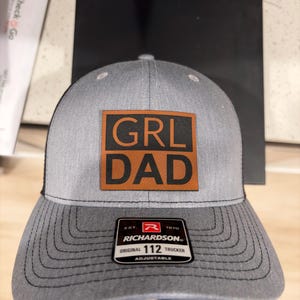 May include: Gray trucker hat with a black mesh back and a curved brim. The front features a brown square patch with the words "GRL DAD" in bold, white letters. The hat includes a Richardson 112 Trucker tag.