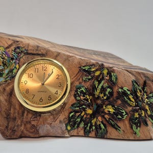 May include: A decorative wooden clock with a gold-colored clock face. The clock is carved from a piece of wood and features painted hummingbird and floral designs in shades of green, yellow, and blue. The clock is approximately 6 inches long.