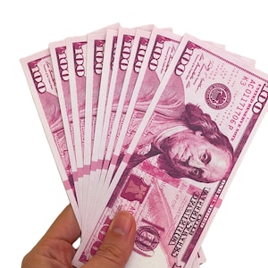 May include: A hand holding a stack of ten pink $100 bills. The bills are fanned out and the text "UNITED STATES OF AMERICA" and "ONE HUNDRED DOLLARS" is visible.