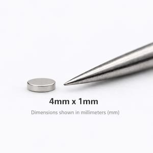 May include: A small, circular, silver-colored metal disc magnet positioned next to a pointed metal tool. The text 4mm x 1mm is printed below the objects, indicating the dimensions shown in millimeters (mm) for this flat, round magnet.