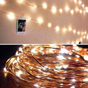 May include: Warm white LED string lights on copper wire, with a photo clipped to the string. The lights are arranged on a wall, with a close-up of the coiled lights below. The lights create a cozy, ambient glow.