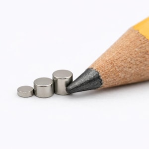 May include: Three small, cylindrical, metallic silver magnets arranged in a row by size, positioned next to the sharpened graphite tip of a yellow wooden pencil to provide a clear visual scale of their miniature dimensions.