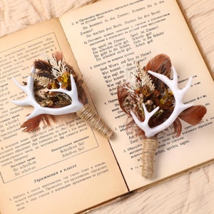 Op de afbeelding: Twee witte hertengewei corsages met gedroogde bloemen en veren. De corsages zijn vastgebonden met touw en liggen op een open boek.