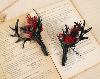 May include: Two black antler boutonnieres with red roses, red berries, and black feathers. The boutonnieres are on a book with German text.