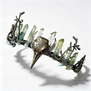 Peut inclure: Une couronne artisanale avec une pièce maîtresse en forme de crâne d'oiseau de couleur bronze et des détails complexes. La couronne est ornée de pointes de cristal vert et d'éléments en forme de branches, créant une esthétique gothique ou fantastique.