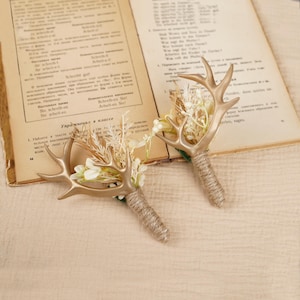 Può includere: Due boutonnière in corna di cervo dorate con fiori secchi bianchi e beige e spago marrone.