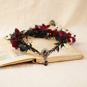 May include: A black and burgundy floral crown with a silver pendant featuring a dark red gemstone. The crown is resting on an open book.