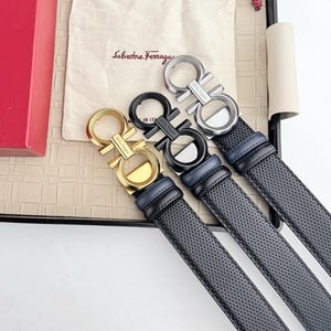 Puede incluir: Tres cinturones de cuero con hebillas doradas, negras y plateadas. Los cinturones tienen una textura perforada y un respaldo de cuero azul. Las hebillas presentan un diseño doble Gancini. Una bolsa beige con el texto "Salvatore Ferragamo" está en el fondo.