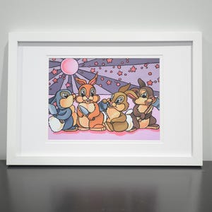May include: A framed art print featuring four cartoon rabbits in a colorful illustration. The artwork has a pink and purple background with a sun and stars. The rabbits are in shades of blue, orange, and brown.