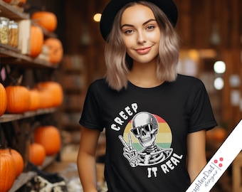 May include: Black t-shirt with a skeleton graphic wearing sunglasses and giving a peace sign. The text "CREEP IT REAL" is above and below the skeleton. The background includes pumpkins and shelves.