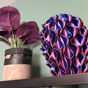 Op de afbeelding: Een decoratieve, abstracte sculptuur met een golvend, gelaagd ontwerp in paarse en blauwe tinten. Daarnaast een potplant met grote, dieppaarse bladeren en een zwarte en beige pot. De items staan op een zwart rek.