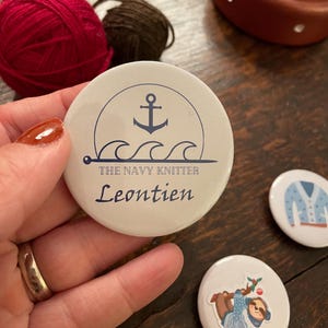 Może przedstawiać: Biały guzik z niebieską kotwicą i falami, pod kotwicą znajduje się napis "THE NAVY KNITTER Leontjen".