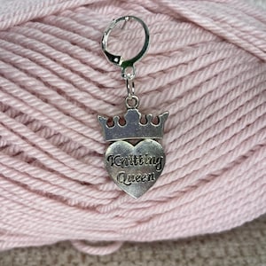 Pode incluir: Marcador de ponto de tricô prateado com um amuleto em forma de coração que diz "Knitting Queen" e uma coroa no topo. O marcador está em uma bola de lã rosa.