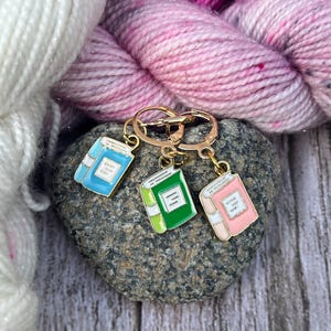 Op de afbeelding: Drie metalen steekmarkeerders in de vorm van miniatuurboeken, elk voorzien van een goudkleurige ringsluiting. De bedels zijn afgewerkt in emaille met lichtblauwe, groene en roze covers, elk met witte rug- en paginadetails aan de voorzijde.