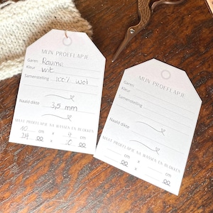 Pode incluir: Duas etiquetas de papel branco com o texto "MIJN PROEFLAPJE" no topo. As etiquetas servem para registar informações de tricô, incluindo o tipo de fio, a cor, o tamanho das agulhas e as medidas do tecido tricotado antes e depois de lavar e bloquear. A primeira etiqueta tem as seguintes informações preenchidas: "Garen: Raumo", "Kleur: wit", "Samenstelling: 100% wol", "Naald dikte: 3,5 mm", "MAAT PROEFLAPIE NA WASSEN EN BLOKKEN: 40 cm x 24 cm x 14 cm x 10 cm".