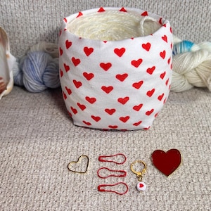 Può includere: Un cestino di filo di tessuto bianco con un motivo a cuori rossi, riempito di filo bianco. Sono mostrati anche segnapunti a forma di cuore dorati e rossi, una spilla a forma di cuore rossa e una conchiglia. Filato e accessori per maglieria.