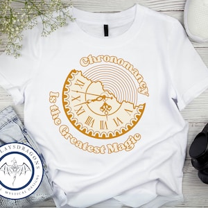 May include: White t-shirt with a gold graphic of a clock with the text "Chronomancy is the Greatest Magic".