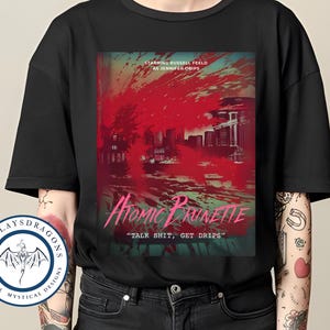 Puede incluir: Camiseta negra con un gráfico de un horizonte de ciudad en rojo y rosa con el texto "Atomic Brunette" y "Talk Shit, Get Drips" en rosa.